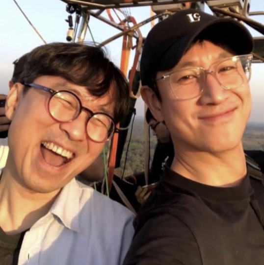 people-Two Asian men-are smiling-while riding in a hot air balloon basket-They appear to be traveling or on vacation-The man on the left is laughing heartily with round glasses and a light-colored shirt, and the man on the right is wearing a black cap and clear-framed glasses-The photo is a selfie taken outdoors, likely during sunrise or sunset, given the warm light.