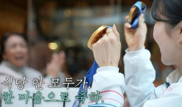 *people-young woman in white uniform-holding-gold medal-excited older woman clapping-Korean text on screen*