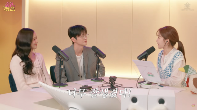 people-Three young people in a podcast studio-The woman on the left has long dark hair and is wearing a light pink/beige cardigan-The man in the center is wearing glasses and a grey-green blazer over a white shirt-The woman on the right has her brown hair in a ponytail and is wearing a white top with a blue patterned vest-Korean text overlay at the bottom says 