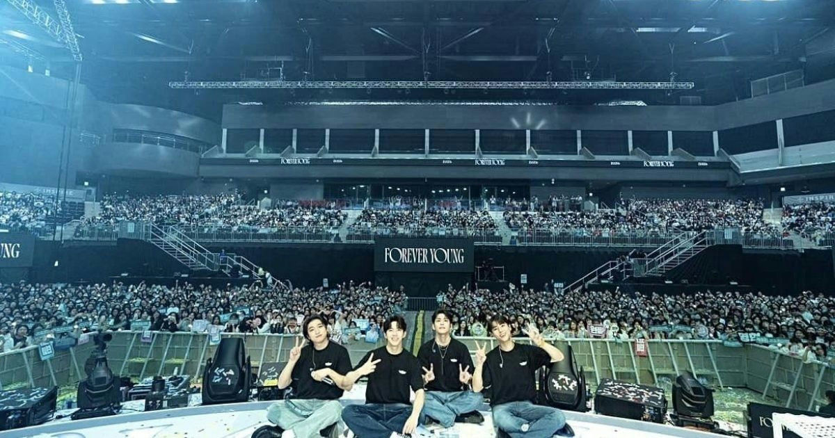 people-Four young men-A-sitting on stage-A-waving peace signs-B-in front of a large crowd-C-at a concert venue