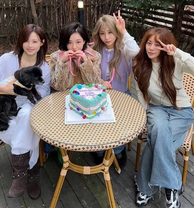 people-Four young women and a small black dog-sitting around a woven outdoor table-with a heart-shaped, light blue and white layered cake on it-The women are dressed in casual but stylish attire, two on the right making peace signs-One woman on the far left is holding the dog-The overall setting appears to be an outdoor patio or garden area-The cake has colorful candy decorations and pink writing on top-The person on the far right is likely Tiffany Young (Hwang Miyoung) from Girls' Generation-The person next to her with blonde hair is likely Kim Hyoyeon, also from Girls' Generation-The person holding the dog is likely Choi Soo-young (Sooyoung) from Girls' Generation-The person in the middle with dark hair is likely Seohyun (Seo Ju-hyun) from Girls' Generation