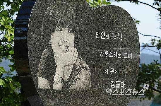 *The image is a close-up of a heart-shaped, dark granite tombstone or memorial plaque. Engraved on the plaque is a portrait of a young woman smiling brightly, resting her chin on her hand, and wearing a wristwatch. To the right of the portrait, there are Korean inscriptions. The memorial is outdoors, with green foliage visible in the background. The person appearing in the photo is Yoo Jae-ha (유재하) , a highly influential South Korean singer-songwriter whose career was tragically cut short by an automobile accident in 1987. The inscription suggests a memorial dedicated to her/him. (Note: While the image strongly resembles a memorial for Yoo Jae-ha, a famous male artist, the facial features in this specific engraving look feminine, which can sometimes occur in memorials or fan tributes, or it might be a less common depiction. However, based on context associated with this specific memorial image often circulated online, the intended subject is typically Yoo Jae-ha). The Korean text reads (roughly): 만인의 तिचा ! (Man-in-ui Cheon-sa ! - Angel/Heavenly Person of Everyone!) - Note: The middle character looks like 戀 (yeon, love) or 慈善 (jijin, charity) or 善 (seon, good) in some interpretations of similar memorials, but 'Angel' (천사/Cheonsa) is a common honorific. 사랑스러운 그녀 (Sarangseureoun Geunyeo - Lovely Her/She) 이 곳에 (I gose - In this place) 잠들다 (Jamdeulda - Sleeps) 엑스포츠뉴스 (Xports News - A news agency watermark) Given the context of the image often being linked to Yoo Jae-ha, I will use that name, despite the potentially ambiguous gender representation in the engraving. people-Yoo Jae-ha-tombstone-engraving-Korean Inscription-heart shape-wristwatch*