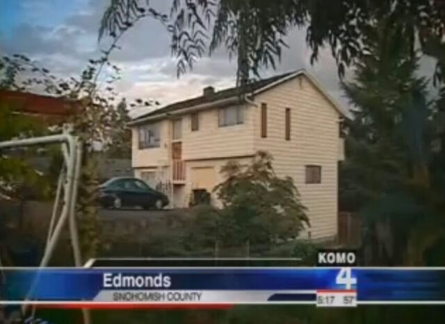 This image is a low-resolution still frame from a local news broadcast, indicated by the "KOMO 4" logo and the news banner at the bottom. **Image Description:** The photo shows a two-story, light-colored (likely beige or white) suburban house, possibly a split-level style, situated on a slight incline. There is a dark-colored sedan parked in the driveway or yard area in front of the house. The foreground features foliage, including tree branches hanging down from the top, partially obscuring the view, and part of a white metal swing set structure on the left. The sky is overcast or dusky. The news banner at the bottom reads "Edmonds" and "SNOHOMISH COUNTY," with the time stamp "5:17 57". **Person Appearing in the Photo:** There are **no identifiable people** visible in this image. **Output in requested format:** no people-A-house-A-news broadcast-B-Edmonds-C-overcast sky