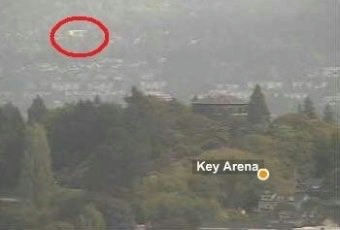The image provided is a landscape photograph featuring a distant view of a cityscape nestled against a hillside, with some prominent green foliage in the foreground. There are labels identifying "Key Arena" in the lower right section. A bright, unidentified object circled in red is visible in the upper left portion of the image, high above the residential area. There are no discernible people appearing in this photo. people-None-A-landscape-A-cityscape-B-Key Arena-C