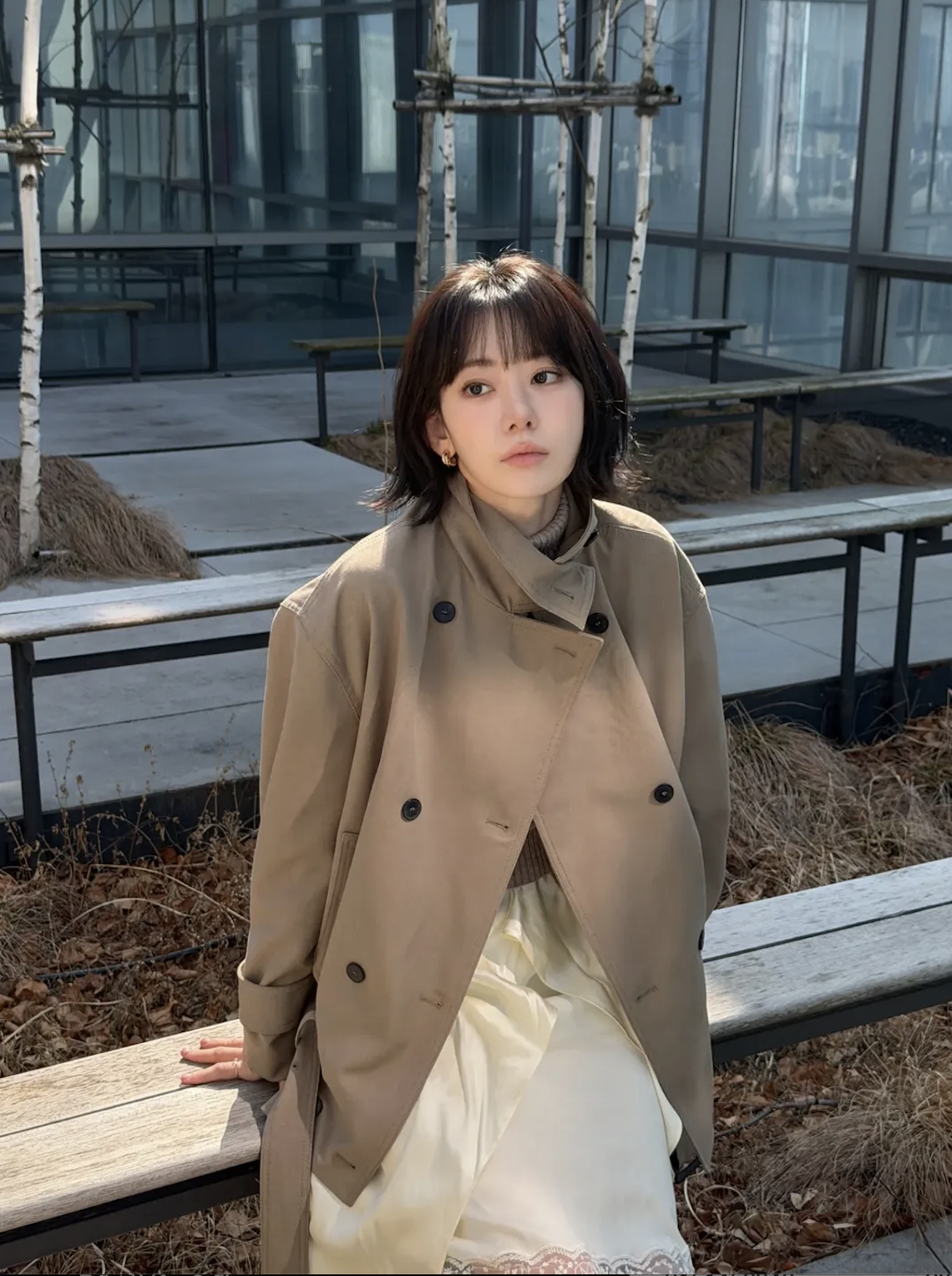 people-Korean actress-A-short dark hair with bangs-A-wearing a beige double-breasted trench coat-B-sitting outdoors on a wooden bench-C-in an urban setting with modern glass buildings and birch trees in the background
