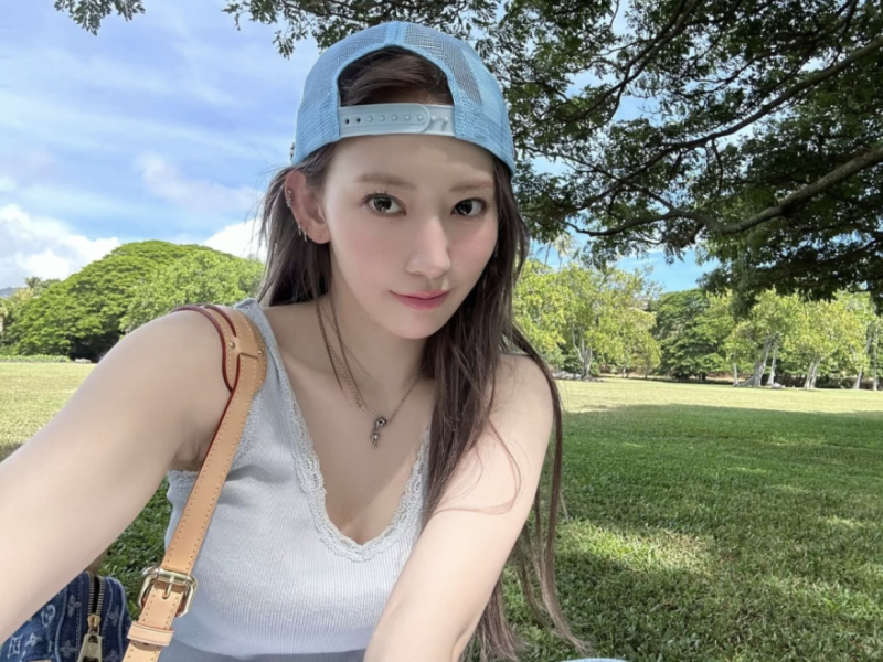people-Kim Chaewon-light blue trucker hat-light blue ribbed tank top with lace trim-tan leather shoulder bag strap-sitting in a grassy park under trees-outdoor daytime selfie