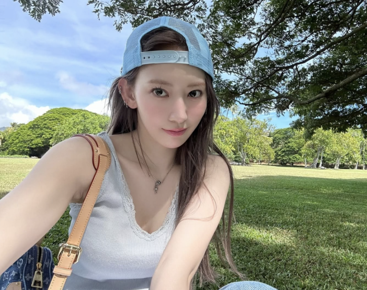 people-Kim Chaewon-light blue trucker hat-light blue ribbed tank top with lace trim-tan leather shoulder bag strap-sitting in a grassy park under trees-outdoor daytime selfie