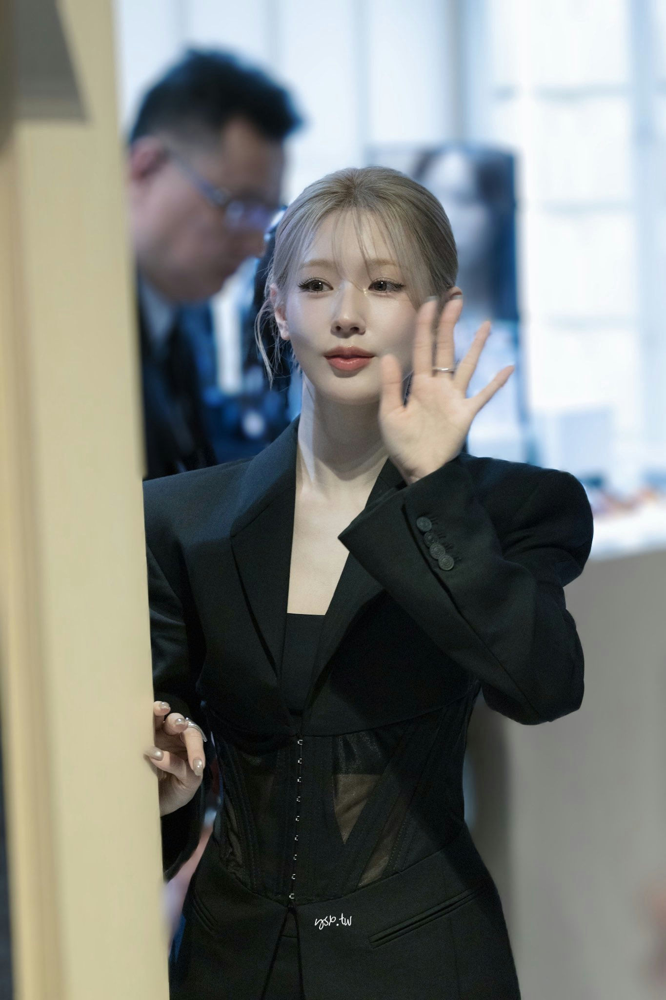 people-Jeon Somi-waving-A-black suit with corset detailing-B-blonde hair styled up with face-framing bangs-C-indoor setting with blurred background figures