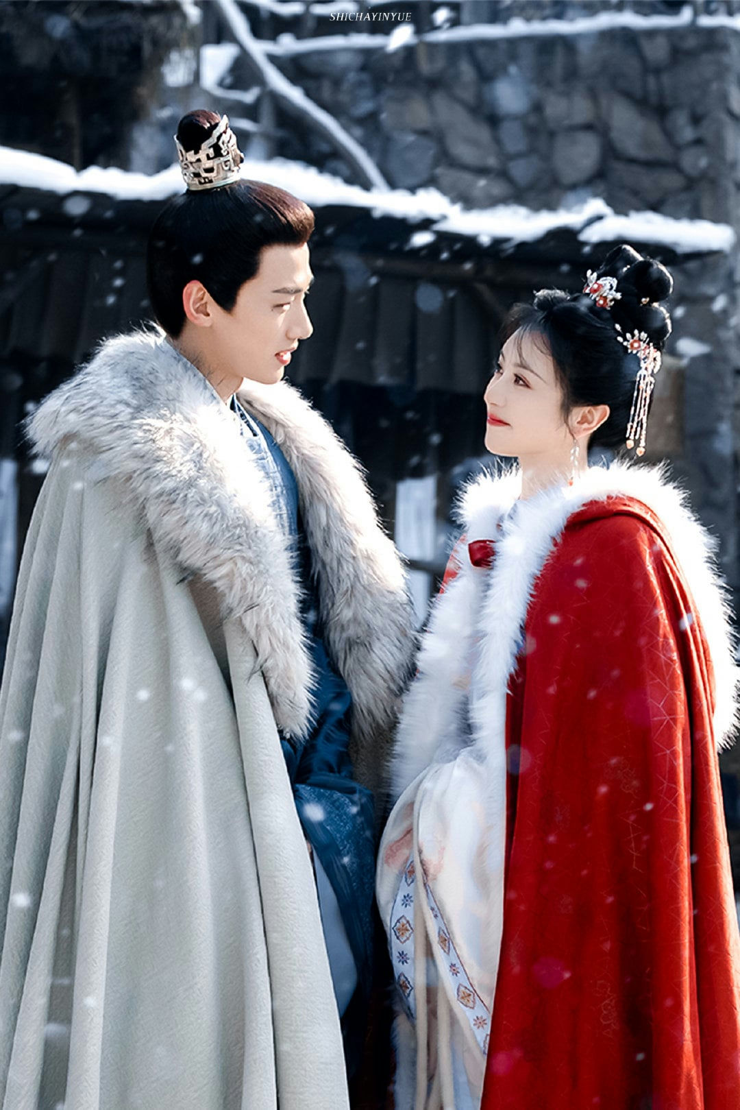 A photograph featuring two individuals, a man and a woman, dressed in elaborate historical or fantasy attire suitable for a cold, snowy setting. The man is on the left, wearing a light grey or white voluminous cape with a thick, light-colored fur collar, layered over a blue garment. He has dark hair styled up and adorned with a small, ornate golden crown or headpiece. The woman is on the right, wearing a striking red cape, also trimmed heavily with white fur, layered over white clothing which has some patterned trim. Her dark hair is elaborately styled in high buns, decorated with ornate hairpins and dangling ornaments. Both individuals are looking toward each other with gentle expressions, against a backdrop suggesting a snowy outdoor environment with dark, possibly wooden or stone structures. The image appears to be a promotional still from a Chinese historical drama (C-drama). The individuals appearing in the photo are actors Yang Zi (the woman) and Xiao Zhan (the man), from the drama **The Longest Promise** (**玉骨遥** / **Yu Gu Yao**). people-Yang Zi-A-Xiao Zhan-A-historical drama-A-snowy scene-A-fur cloaks-A-Chinese costume drama