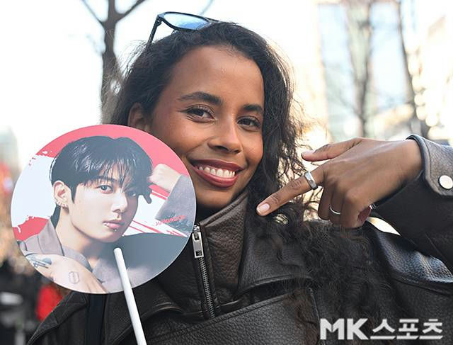 people-Woman smiling outdoors holding a fan-Woman holding a circular fan-Fan features a portrait of Jungkook from BTS-Woman wearing a leather jacket and rings