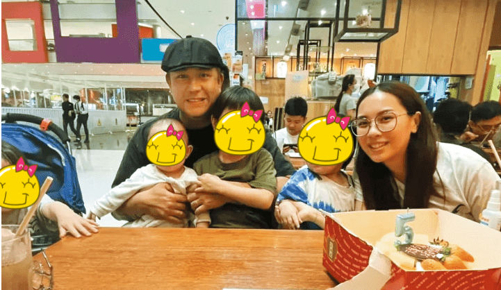 *people-Man with cap holding a child-Woman with glasses smiling-Children with censored faces-Birthday celebration with cake-Family gathering in a public area (possibly a mall or restaurant)*