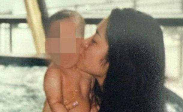 A photo showing a woman with long dark hair kissing a baby on the cheek. The baby's face is pixelated for privacy. The image quality is somewhat faded or aged. The woman is likely the mother, given the affectionate pose. Without further context or identification, the person cannot be named. people-woman-A-baby-B-kissing-C-affection