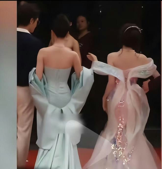 people-Two women in elaborate gowns, viewed from the back-Woman in light blue strapless gown with voluminous sleeves-Woman in white sheer gown with open back and embellishments-Man in dark blazer and light pants-Woman in dark top blurred in the background