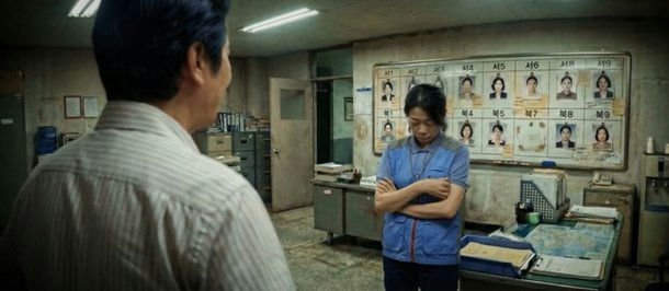 people-South Korean crime drama-A-police office setting-A-woman in blue vest standing with arms crossed-A-man in striped shirt facing away-A-mugshot board with Korean text and female faces
